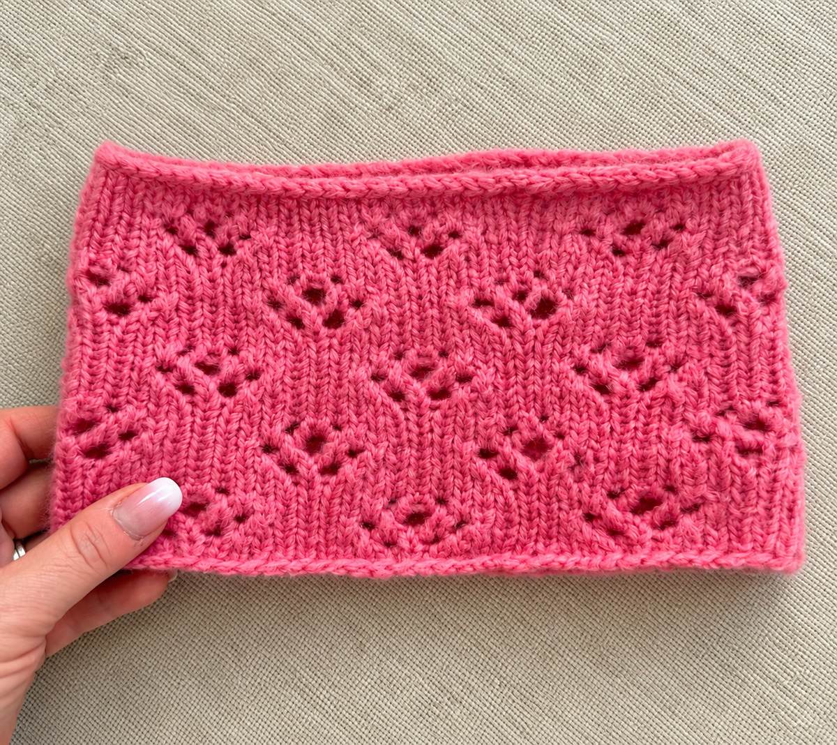 halsen lagt flatt: Rosa Rosella Cowl fra Leneknit lagt flatt på lys bakgrunn, med detaljer av hullmønster og i-cordkant.
